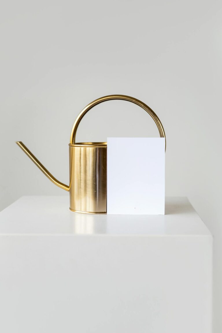 Minimalist gold watering can beside a blank white card on a white plinth, perfect for mockup designs.